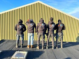 Vijf personen in werkkleding staan met hun rug naar de camera toe voor een groen gebouw.
