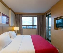 Quarto de hotel com cama de casal, roupa de cama vermelha, janela grande e TV na parede.