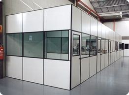 Sala modular em um galpão com paredes brancas e janelas de vidro, piso cinza e teto alto.