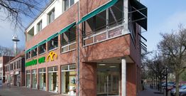 Eckgebäude aus rotem Backstein mit großen Fenstern und einem grünen Ladenschild in einer Straße.