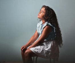 Niña sentada con vestido azul claro, mirando hacia arriba, fondo gris.