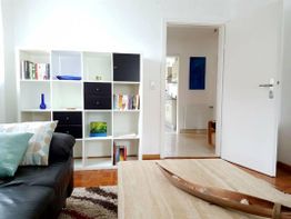 Helles Wohnzimmer mit Regal, Sofa und Tisch. Tür öffnet sich zur Küche.