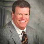 Smiling man in a gray suit with a patterned tie, against a gray backdrop.