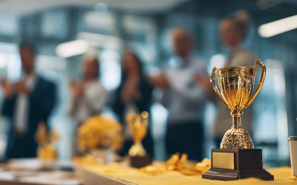 Pokal, Medaille und Trophäe auf einem Tisch, im Hintergrund ein applaudierendes Team, symbolisiert Wertschätzung und Motivation im Unternehmen.