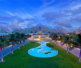 Complejo hotelero iluminado, con fuente y palmeras al atardecer bajo un cielo azul.