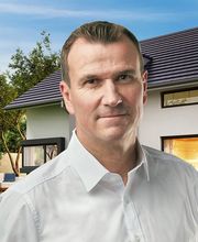 Mann in weißem Hemd vor modernem Haus mit Garten und Pool im Hintergrund.