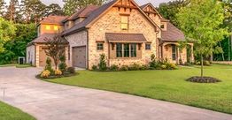 A large brick house with a manicured lawn and driveway surrounded by trees.