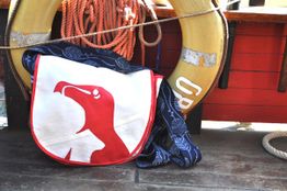 Umhängetasche mit rotem Vogelmuster vor Rettungsring und Seilen auf hölzernem Bootsdeck.