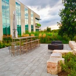 Modern patio with tables, chairs, stone benches, greenery, and a glass building under cloudy skies.