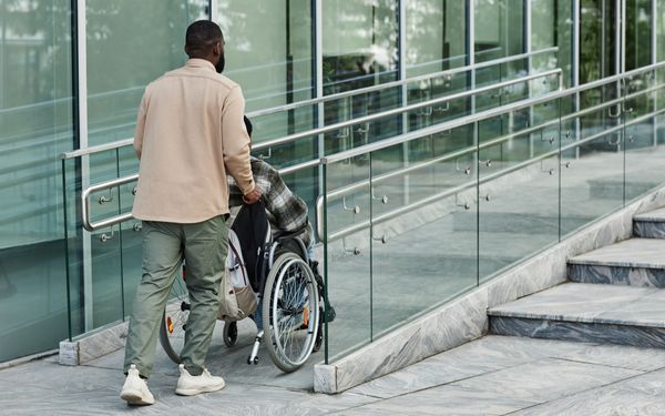 Eine Person schiebt einen Rollstuhlnutzer über eine barrierefreie Rampe neben einer Treppe in ein modernes Gebäude.