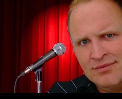 Mann neben Mikrofon vor roten Vorhängen, mit ernstem Gesichtsausdruck.