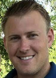 A man smiling outdoors, with short hair and a leafy background.