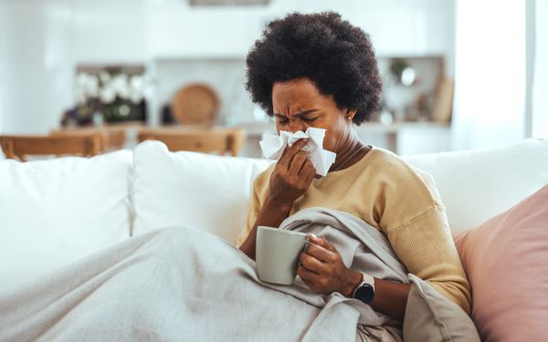 Frau sitzt erkältet auf dem Sofa, hält eine Tasse in der Hand und putzt sich mit einem Taschentuch die Nase.