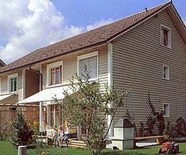 Zweistöckiges Haus mit Garten, Sonnenschutz und Holzwandverkleidung, blauer Himmel im Hintergrund.