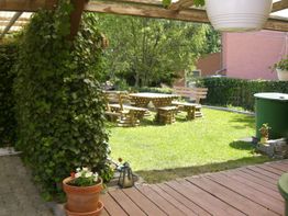 Garten mit Holzbänken, Tisch, Pflanzen und einem grünen Regenfass unter einem überdachten Bereich.