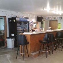 Empty bar interior with black stools, a stocked fridge, and drinks on the counter.