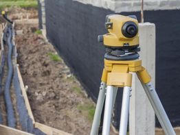 Surveying equipment on tripod near construction site with black foundation wall.