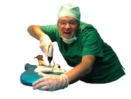 Mann in OP-Kleidung repariert E-Gitarre mit Bohrmaschine, lächelnd.