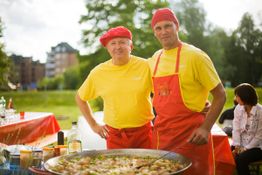 Zwei Männer in gelben T-Shirts und roten Schürzen kochen Paella im Freien auf einem Fest.