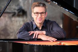 Eine Person mit Brille lehnt lächelnd an einem Flügel.