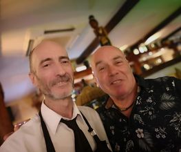 Dos hombres sonríen en un bar bien iluminado. Uno viste camisa formal, el otro camisa floral.