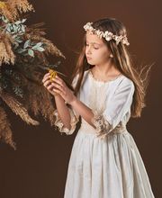 Niña con corona de flores y vestido blanco, observando un ramo de hojas secas y flores.