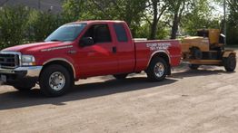 Red truck towing stump removal equipment on a dirt road, surrounded by trees.