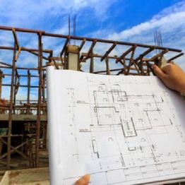 Person holding building plans in front of a construction site under a blue sky.