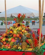 Obsttisch am Strand mit Zelthintergrund, dekoriert mit Ananas und roten Blumen.