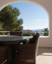 Terraza con mesa y sillas de mimbre, vista al mar y árboles al fondo.
