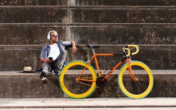 Man lehnt lässig auf einer Treppe, hört dabei Musik über Kopfhörer. Vor ihm steht sein Fahrrad in knalligen Farben.