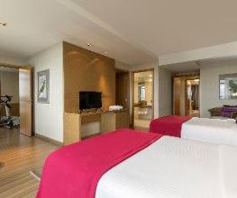 Quarto de hotel com duas camas, TV, mesa e entrada para banheiro e academia ao fundo.