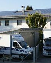 Transporter und LKWs stehen vor einem Gebäude mit Solarpanels, umgeben von Bäumen.