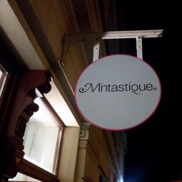 Rundes Schild mit dem Text "Mintastique" hängt an einer Hauswand bei Nacht.