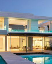 Moderne Villa mit Glasfronten, beleuchtet bei Dämmerung, mit Pool und Palmen im Garten.