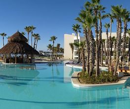 Piscina exterior con palmeras y cabaña de paja, junto a hotel moderno bajo un cielo despejado.