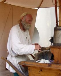 Ein Mann mit Bart arbeitet in historischer Kleidung an einem Schleifstein unter einem Zelt.