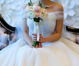 Sposa con abito bianco decorato, seduta su un divano, tiene un bouquet di fiori rosa e bianchi.