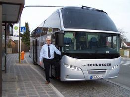 Mann steht lächelnd neben silbernem Reisebus mit der Aufschrift "Dr. Schlosser" an Haltestelle.