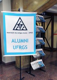 Banner da Alumni UFRGS ao lado de um suporte de revistas em um ambiente interno.