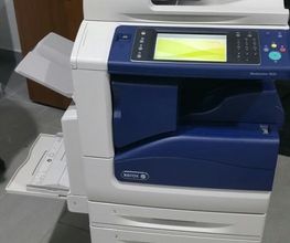 Stampante multifunzione bianca e blu in un ufficio, con display acceso e vassoio aperto.