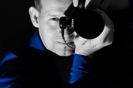 Ein Mann in blauem Hemd fotografiert mit einer Kamera aus der Nähe. Gesicht nur teils sichtbar.