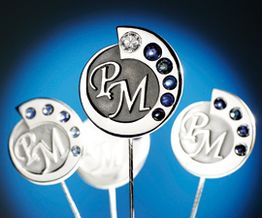 Runde Broschen mit PM-Logo und blauen Edelsteinen, vor blauem Hintergrund arrangiert.