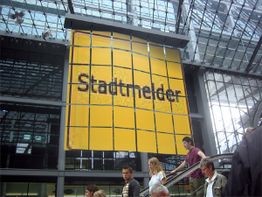 Personen auf Rolltreppe vor einem großen gelben Schild mit Aufschrift "Stadtmelder" im Gebäude.