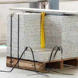 Pallet of stacked concrete slabs secured with straps, featuring a yellow strap on a construction site.