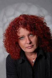 Frau mit lockigem, rotem Haar und schwarzer Kleidung vor grauem Hintergrund.