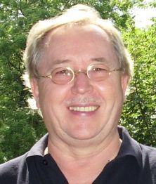 Ein lächelnder Mann mit Brille vor einem Hintergrund aus grünen Bäumen.