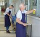 Two janitors wearing aprons clean windows and stack chairs in a hallway.