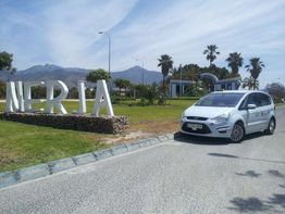 Coche blanco cerca de letras grandes de "NERJA" en una rotonda con montañas al fondo.
