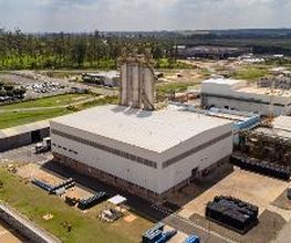 Fábrica em área industrial com vegetação ao fundo e céu parcialmente nublado.